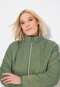 Giacca trapuntata verde con colletto alto e chiusura con zip. Presenta un motivo testurizzato e un logo in evidenza sulla manica.