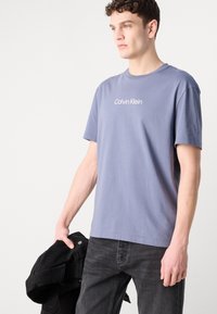 Gaiši zila kokvilnas T-kreklā, apaļš kakla izgriezums, īsas piedurknes, ar "Calvin Klein" logo izdrukātu uz krūtīm. Savienots ar tumšiem džinsiem un melnu jaku.