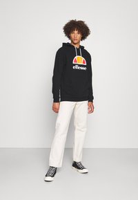 Sudadera negra con un colorido logo gráfico, que presenta un bolsillo frontal y capucha con cordón, combinada con pantalones beige y zapatillas altas negras.