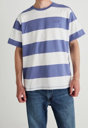 Man die een blauwe en witte horizontaal gestreepte Levi’s T-shirt en blauwe spijkerbroek draagt, staand tegen een effen achtergrond.