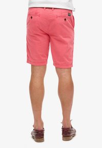 Pantalones cortos de color rosa claro hechos de algodón, con bolsillos traseros con cierres de botón y un corte recto, combinados con zapatos marrones de tipo slip-on.