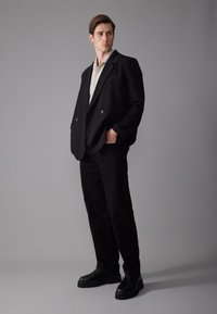 Blazer noir à double boutonnage sur une chemise beige clair, associé à un pantalon noir et des chaussures noires imposantes, sur fond gris.