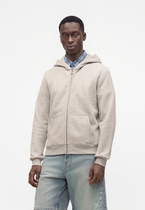 Mand iført en lys beige hættetrøje med lynlås over en blå ternet skjorte og falmede denimbukser med stjerne-patches, stående mod en ensfarvet baggrund.