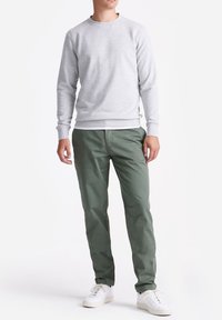 Man in een lichtgrijze sweatshirts, witte onderhemden, olijfgroene broek en witte sneakers staat tegen een effen witte achtergrond.