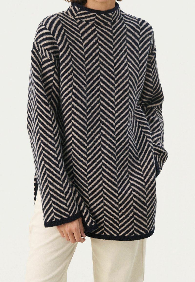 Personne portant un pull à manches longues bleu marine et beige à rayures chevrons avec un col montant et un pantalon clair, debout devant un fond blanc.