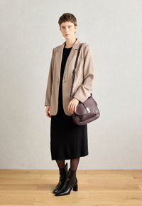 Blazer beige sur une robe noire, associé à des bottines noires. Le mannequin tient un sac en cuir violet foncé avec un accent argenté.
