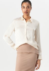 Long-sleeve cream blouse in lightweight fabric, featuring a subtle patterned texture, button-up front, and collar, paired with a fitted skirt.