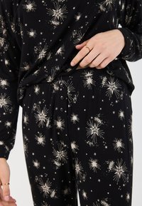 Ensemble de vêtements de détente noir avec un motif étoilé en crème. Le haut est ample avec des manches longues ; le pantalon possède un détail en bouton et une texture douce.