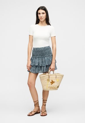 Γυναίκα με λευκό κοντομάνικο τοπ, μπλε μίνι φούστα με βολάν και λουλούδια, καφέ δερμάτινα σανδάλια με κορδόνια, κρατώντας ψάθινη τσάντα με τροπική κεντητή διακόσμηση.