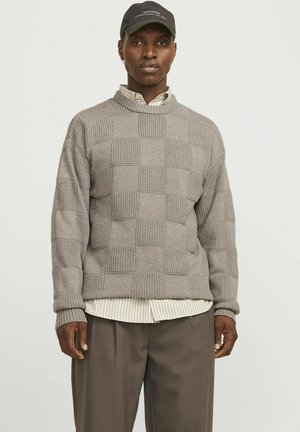 Pull en tricot marron clair avec un motif à carreaux, présentant un col montant et des poignets côtelés, superposé à une chemise à col à rayures.