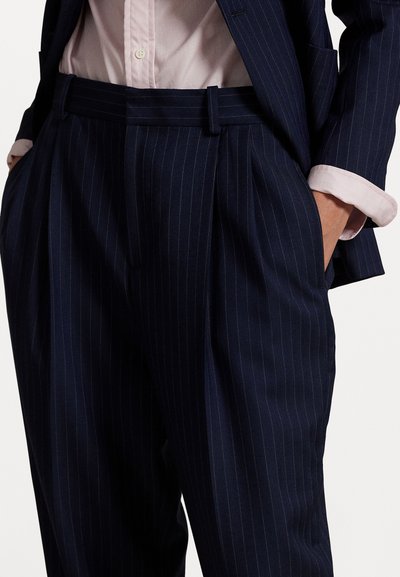 Navy blue pinstriped suit pants with pleats, side pockets, and a textured fabric; worn with a light pink shirt's cuff visible.