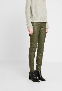 Olivgröna smalnande byxor i konstläder i kombination med en beige stickad tröja och svarta klackade ankelboots med ribbat manschetter.