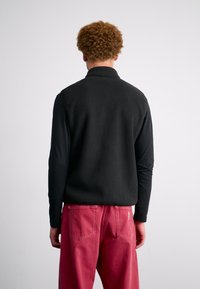 Melnā vilnas veste ar teksturētu virsmu, valkāta virs garām piedurknēm melnas krāsas krekla. Apvienota ar spilgti sarkanām plašām biksēm.