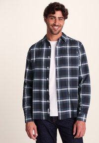 Men's long-sleeve plaid shirt featuring navy and light blue tones, button front, chest pocket, and soft fabric texture. Worn over a white t-shirt.