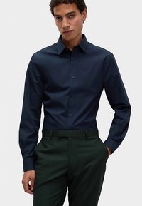 Marineblaue Langarmbluse aus glattem Stoff; verfügt über einen Standardkragen und eine durchgehende Knopfleiste. Kombiniert mit dunkelgrünen Hosen.