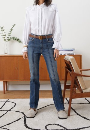 Persoon die een wit blouse draagt met een gerimpelde kraag en manchetten, blauwe spijkerbroek met voorste naden, bruine riem en beige instapschoenen, staand binnen.