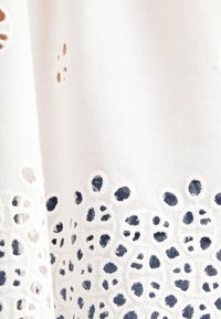 Indumento in tessuto bianco caratterizzato da intricati modelli a taglio lungo il bordo inferiore, con una superficie morbida e testurizzata e una leggera lucentezza.