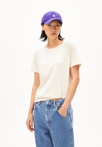 Beige Baumwoll-T-Shirt mit kurzen Ärmeln, kombiniert mit hellblauen Jeans. Das Model trägt eine lila Mütze mit einem Logo.
