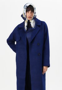 Navy blue wool coat with a wide collar, double-breasted buttons, and a chest pocket. Accompanied by a patterned silk scarf.