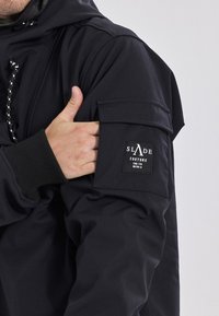 Zwarte hoodiejack met een gestructureerde afwerking, voorzien van een patchzak op de mouw en een logolabel. Detail van de trekkoordkap zichtbaar.