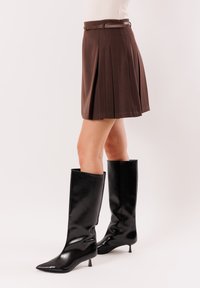 Donna che indossa una gonna marrone plissettata con una cintura sottile e stivali al ginocchio neri con punta appuntita e tacchi bassi, in piedi su uno sfondo neutro.