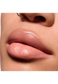 Primo piano di labbra piene con un colore nude opaco e morbido e una leggera lucentezza, che mostrano una consistenza liscia e linee sottili. Un orecchino a cerchio argentato visibile.
