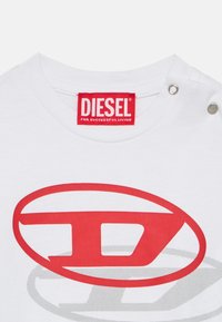 Λευκό κολάρο πουκάμισου με κόκκινη ετικέτα της μάρκας Diesel και δύο μεταλλικά κουμπιά τύπου snap, με μεγάλο κόκκινο και γκρι στυλιζαρισμένο λογότυπο "D" πάνω στο ύφασμα.