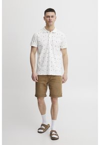 Weißes Polohemd mit bunten Mustern, kombiniert mit hellbraunen, hochgekrempelten Shorts, weißen Socken und braunen Sandalen. Das Modell steht aufrecht.
