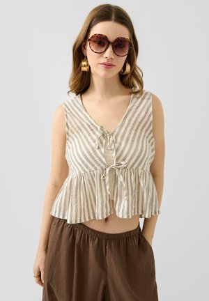 Femme portant de grandes lunettes de soleil écaille, des boucles d'oreilles dorées en anneau, un top sans manches beige rayé avec un lien devant et un pantalon large marron.