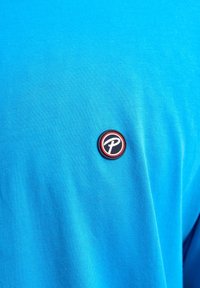 Tricou din bumbac albastru, decorat cu un logo brodat rotund, în culorile negru și roșu, având o literă albă în centrul său, expus pe partea stângă sus.