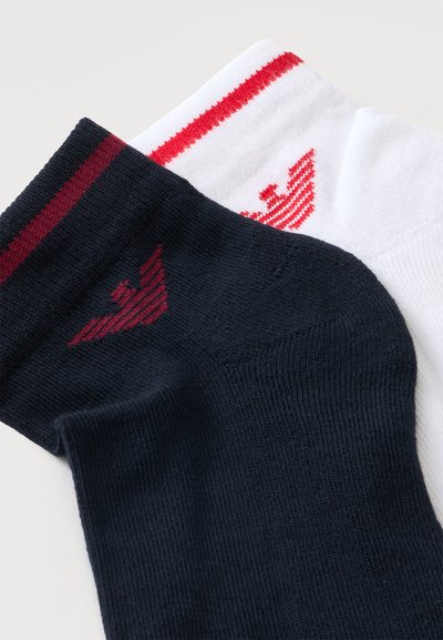 Deux chaussettes courtes pliées, une blanche avec des rayures rouges et un logo, une noire avec des rayures bordeaux et un logo, sur fond blanc.