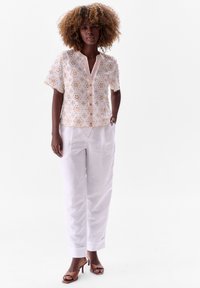 Witte bloementextuur blouse met korte mouwen en een knoopsluiting aan de voorkant, gecombineerd met losse witte broek en bruine sleehakken.