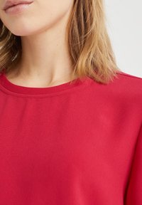 Top leggero rosso in tessuto con scollo rotondo e superficie liscia. Dettaglio ravvicinato della zona del collo e della spalla, che mostra sottili dettagli di cucitura.