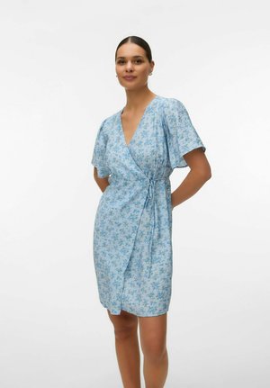 Femme debout, les mains derrière le dos, portant une robe portefeuille bleu clair à motifs floraux avec des manches courtes, sur fond blanc.