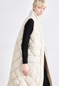 Colete bege acolchoado sem mangas com colarinho alto, apresentando botões de pressão, bolsos laterais e um design longo e fluido. Blusa de mangas longas preta por baixo.