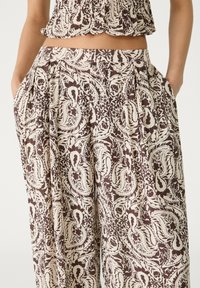 Hoge taille losse broek en bijpassende cropped top met een bruine en crèmekleurige bloemen paisleyprint, model met de handen in de zakken.
