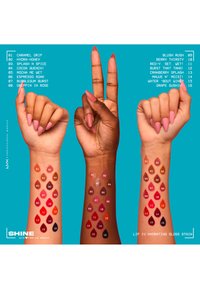 Trois bras levés avec de longs ongles roses affichent 16 nuances de taches de gloss à lèvres sur les avant-bras, étiquetées avec des noms et des numéros sur un fond bleu.