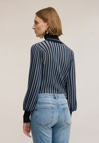 Modrá a černá pruhovaná blůza s vysokým výstřihem a nabíranými rukávy, kombinovaná se světle modrými džíny. Materiál má mírný lesk.