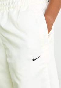 Gaiši beidži Nike treniņbikses ar elastīgu jostu, sānu kabatām un melnu logotipa detaļu kreisajā augšstilbā. Gluda auduma tekstūra.