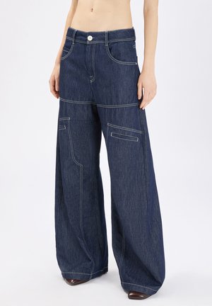 Persona che indossa pantaloni di jeans a vita alta, gamba larga, di colore blu scuro con cuciture bianche a contrasto e tasche con patta, in piedi davanti a uno sfondo semplice.