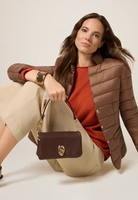 Doudoune marron, pull rouille, pantalon beige à large jambes. Le mannequin tient un petit sac à main en cuir marron avec des garnitures en or et des accents en chaîne.