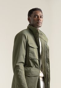 Chaqueta verde caqui con un cuello alto, cierre frontal y botones a presión. Presenta dos bolsillos en el pecho y un tejido texturizado. Ajuste entallado.