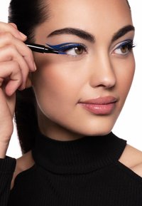 Eyeliner blu applicato sulla palpebra inferiore, pelle liscia della modella, colore neutro per le labbra e dolcevita a coste nero. Sfondo chiaro e luminoso.
