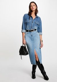 Denim overhemd met knopen, twee borstzakken en opgerolde mouwen, gecombineerd met een licht denim rokje met een split aan de voorkant. Zwarte handtas en laarzen.