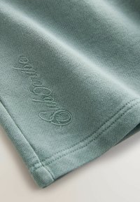 Jasnozielony materiał na sweter z gładką teksturą, z wyszywanym logo oraz obszyciem na dole. Prosty i schludny design.