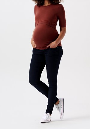 Těhotenský top v barvě rez, přiléhavý s krátkými rukávy, v kombinaci s tmavě modrými skinny džíny a bílými teniskami s červenými akcenty.