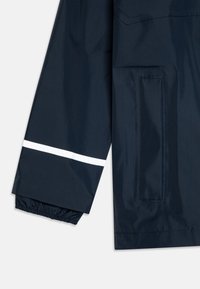 Marine vanntett jakke med erme i elastisk mansjett, med en hvit reflekterende stripe og en sidelomme med klaff. Glatt, glansfull tekstur.