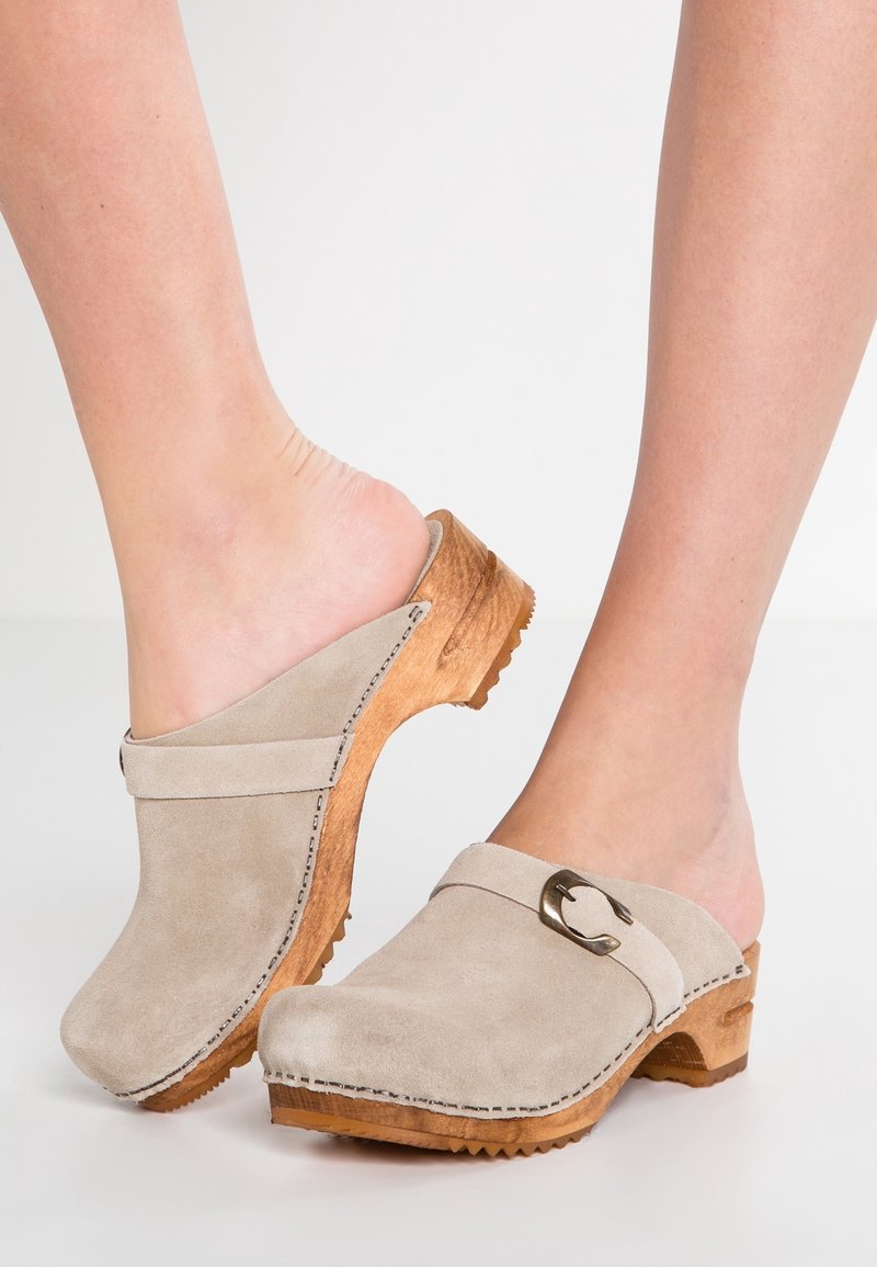 Pieds portant des sabots beige en daim à enfiler avec des talons en bois et une boucle métallique sur une chaussure, sur fond blanc uni.