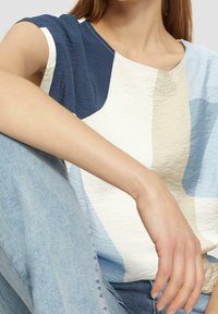 Kortærmet top med tekstureret stof i et abstrakt mønster i blå, hvid og beige. Bæres med lyseblå jeans, delvist synlig hånd.