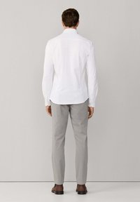 Mężczyzna stoi przodem do ściany, ubrany w biały koszulę z długim rękawem, jasnoszare spodnie i brązowe eleganckie buty.